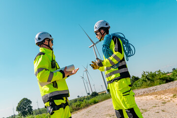 Two workers in bright safety gear discuss wind turbine maintenance at an outdoor site under clear skies