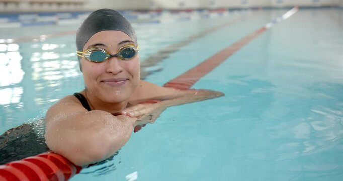 Smiling female swimmer resting at pool edge, wearing swim cap and goggles, copy space