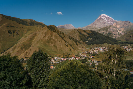 The snowy peak of the mountain, at the foot of the village. Mount Kazbek in Georgia in August. The mountain rises above the city. The village of Stepantsminda. Sunrise in the mountains