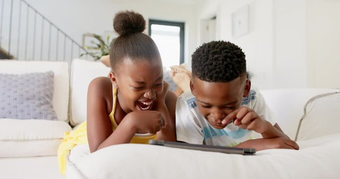 African American siblings lying on couch, watching and laughing at tablet screen