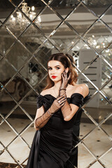 woman in an elegant black dress with open shoulders. She stands in front of a mirrored wall that is made up of many diamond-shaped mirror tiles.