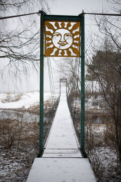 Snow covered hanging bridge over a small river in winter, decorated with yellow metal sun symbol cut out.