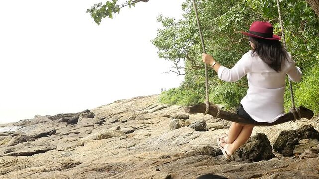 Serene woman in a red hat swings on a tree rope overlooking rocky terrain by the sea, surrounded by lush greenery and nature's tranquility