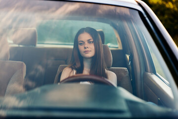 Fototapeta premium Young woman with long brown hair sitting in a car, conveying a sense of contemplation and calmness, surrounded by nature and soft sunlight.