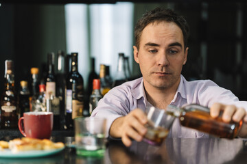 young man drinks whiskey from a glass in a bar on a dark background. A businessman with a bottle in his hands is relaxing in a pub with alcohol after work.
