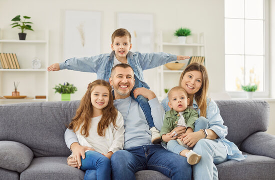 Portrait of smiling young happy family with three kids sitting on sofa at home and looking cheerful at camera together. Mother and father spending time with children. Family time and leisure concept.