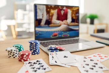 Laptop placed on wooden desk displaying online poker game with dealer, surrounded by colorful poker chips and cards. Screen showing ongoing game, virtual gambling and online gaming experience.