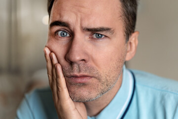 young guy with a sad emotion on her face holding his hand over the patient's jaw on a grey background. Portrait of a man in painful expression with a toothache.