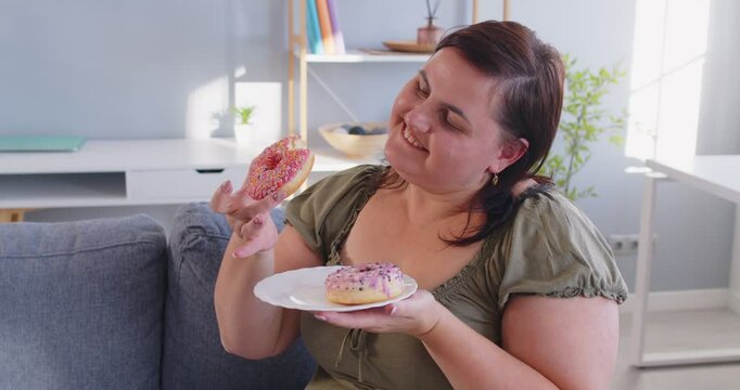 Happy smiling overweight woman enjoying donuts while sitting on sofa at home. Satisfied fat female indulging in sweet treats, showing carefree attitude, love for desserts and unhealthy eating habits.