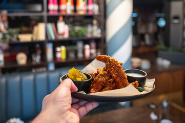 A hand holds a black plate with crispy, sauced chicken wings, accompanied by pickles and dipping sauces. The background features a stylish eatery with colorful decor elements.