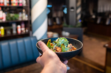 A close-up of a hand holding a black bowl filled with a vibrant salad, featuring greens and...