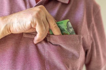Close-up on a man's hand taking a one hundred Australian dollar note into his shirt's pocket. Concept illustration for bribing, corruption or cash payment.