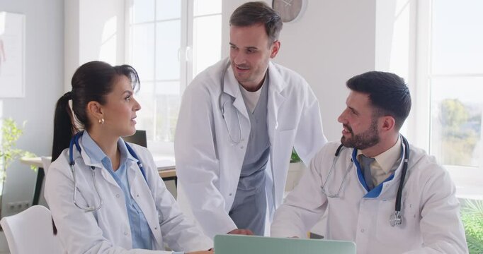 Team three happy smiling doctors in white medical coats working in office, using laptop computer, marking successful development of new effective cure or diagnostic method, giving each other high five