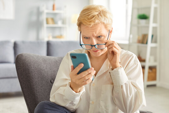 Senior woman sitting on armchair at home reading bad news in internet on phone, feeling sad and upset. Leisure time online is marked by disappointment and concern in a calm environment. - Powered by Adobe