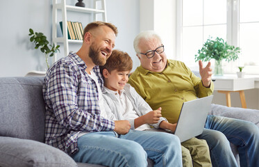 Happy multi generation male family sitting on sofa, using laptop together for online entertainment at home. Father, son and grandfather surfing internet, watching funny videos in relaxed atmosphere.
