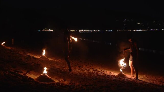 A performer spins fire poi on a sandy beach at night, with distant shoreline lights adding depth to the coastal scene in Thailand.