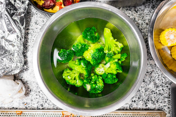 Fresh broccoli soaking in water, surrounded by kitchen utensils and ingredients. The stainless steel bowl contrasts with the colorful vegetables, highlighting a healthy meal preparation scene.