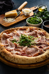 Pizza with cured ham, parmesan cheese and arugula on black table