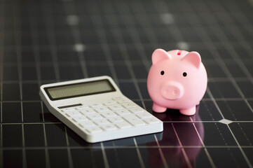Closeup white calculator and pink piggy bank on modern black photovoltaic solar panel for sunlight...