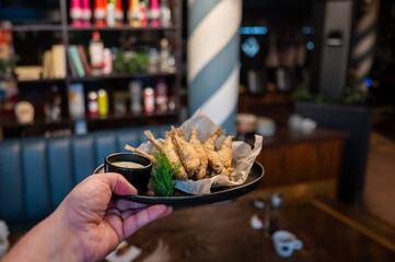 A hand presents a plate of freshly fried fish garnished with herbs and served with a small bowl of dipping sauce, set in a cozy restaurant ambiance.