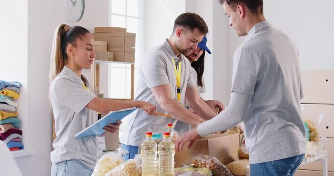 Volunteers group sorting donations of foodstuffs in cardboard boxes for charity food drive. Young smiling people volunteering in food bank working in charitable foundation together. 4k video.