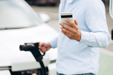 Close up of man with white electric car on the charge station, holding smartphone outdoors
