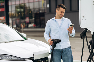 Young smiling handsome man standing at the charging station, looking at phone screen