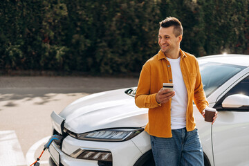 Smiling young Caucasian man charging electric car holding coffee and smartphone outdoors