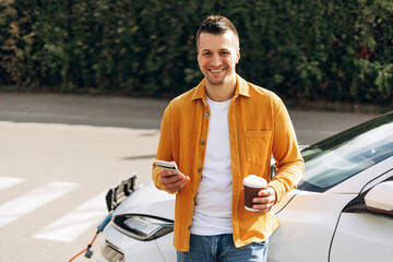 Happy young man with coffee and smartphone having stop at charging station in city