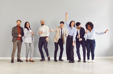 Obraz premium Group portrait of joyful smiling multiracial business team standing in row against grey wall and having fun at end of work day. Happy cheerful company employees dancing together, celebrating weekend.