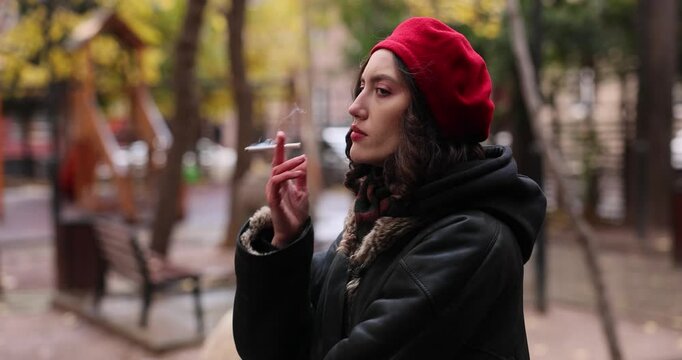 Young woman in red beret smokes cigarette gesturing in autumn park setting. Stylish brunette lady smoker spends time in city garden slow motion