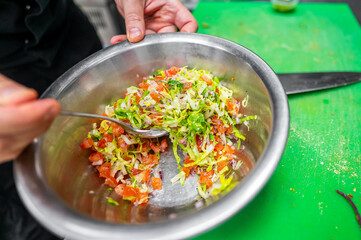A close-up of a chef mixing a fresh salad in a stainless steel bowl. Vibrant ingredients include chopped tomatoes, lettuce, and onions on a green cutting board.