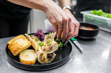 A chef carefully garnishes a plate of bone marrow with fresh herbs and condiments, accompanied by crispy toasted bread. A vibrant kitchen setting enhances the culinary atmosphere.