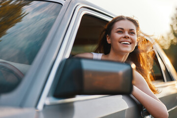 Young woman with long hair enjoying a road trip, smiling joyfully while leaning out of a car window...