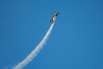 Golden stunt aerobatic plane performing stunts in blue sky with smoke trail. Concept of speed, difficulty, precision, risk