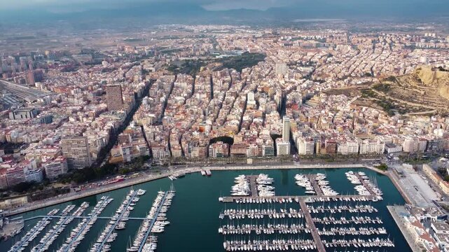 Spectacular aerial perspective of Alicante city. Panoramic aerial view of city center and promenade. View of Port of Alicante. Boats and yachts docked. Famous travel destination. Sunset. Backwards.