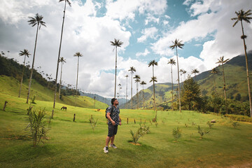 A man stands in Colombia's Cocora Valley, surrounded by lush green hills and towering wax palms, under a dramatic cloudy sky. Perfect for nature, adventure, and travel imagery