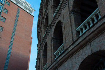 Brick facade of historic bullring las arenas contrasting with modern red building, glass windows reflecting blue sky in barcelona urban landscape