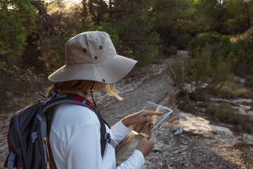 Female biologist wearing a beige hat and carrying a backpack is checking her position on a map...