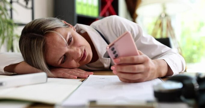 A stressed woman is laying on her cluttered desk, staring at her phone while surrounded by numerous documents