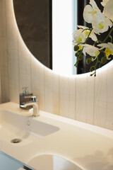 White orchid decorates a modern bathroom featuring a sink, faucet, and a circular mirror with built-in lighting, enhancing the elegant ambiance