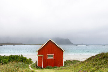 Small old and dilapidatedred (fisherman) cabin (rorbu) along shore of Ramberg Beach, Ramberg, Norway on the Lofoten island chain archipelago with mountains covered in heavy clouds in the background