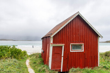 Small old and dilapidatedred (fisherman) cabin (rorbu) along shore of Ramberg Beach, Ramberg, Norway on the Lofoten island chain archipelago with mountains covered in heavy clouds in the background