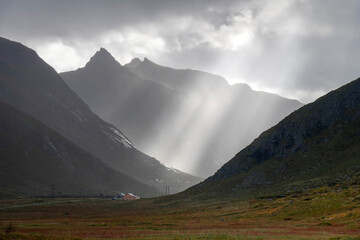 Bright sun rays on lone yellow farmhouse in valley surrounded by mountains and dramatic cloudscape with dark and light clouds near Ramberg in Norway on Lofoten island chain archipelago