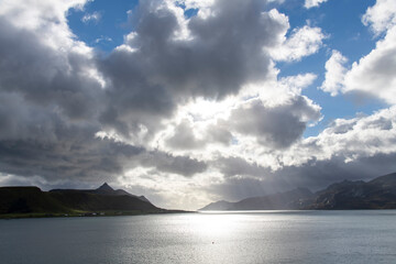 Dramatic cloudscape with dark and light clouds and sun shining bright against silhouette of mountains along one of the fjords near Leknes in Norway on Lofoten island chain archipelago