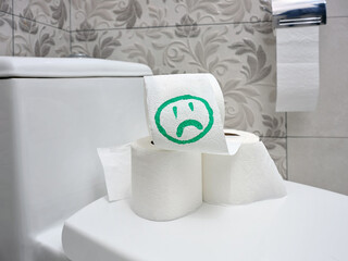 A several rolls of toilet paper with a sad smiley face drawn on it lies on the toilet lid in the apartment's bathroom.