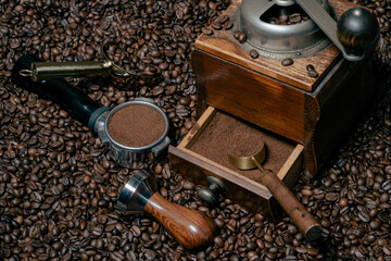 Rustic coffee grinder with a measuring spoon stands  with a tamper and a coffee portafilter filled with freshly ground coffee on a bed of many coffee beans