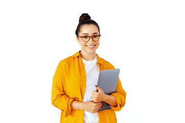 Friendly caucasian or arabian woman in a yellow shirt with glasses, manager or teacher, holding a closed laptop, standing against a white isolated background, looks at camera, smiling confidently