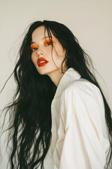 Pretty Asian woman with long black hair, wearing a white oversized shirt, bright orange eyeshadow, and red lipstick, posing in a studio with a minimal beige background in a long shot.