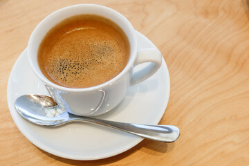 close up of hot black coffee with foam on top in a white cup with wooden table and tea spoon